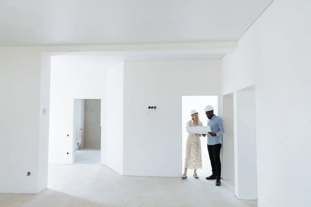 Two people in white hard hats review a plan inside an unfinished home