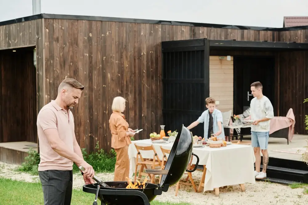 Man grilling on a backyard barbecue while family sets the table
