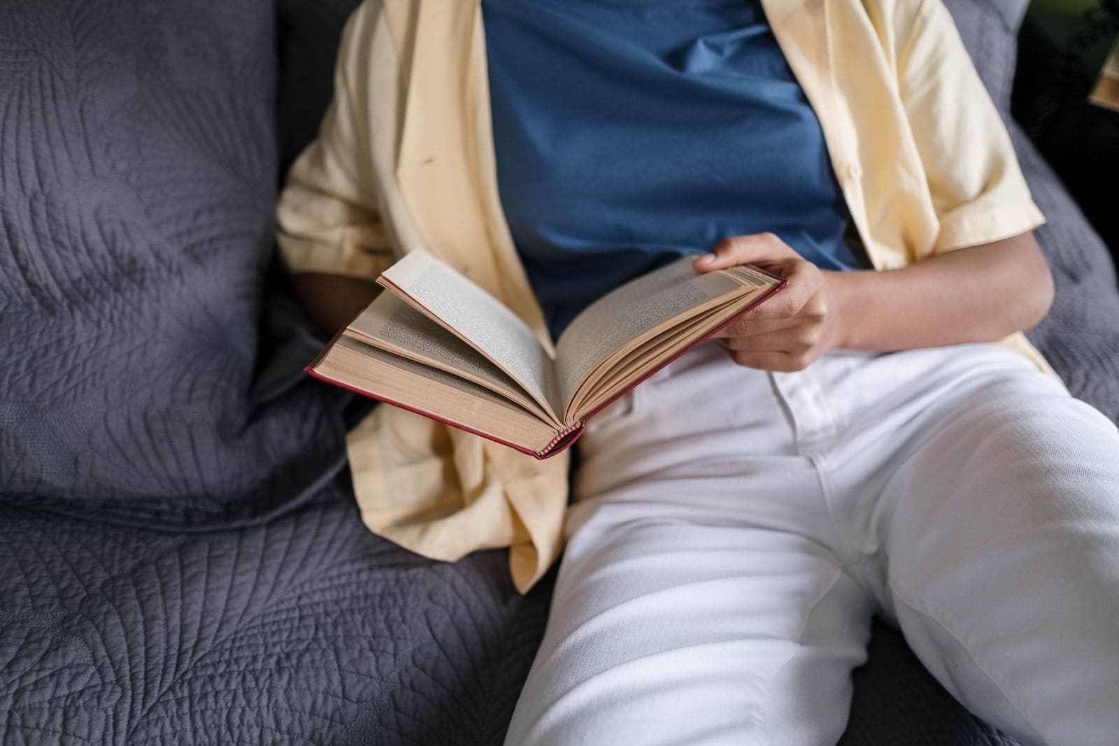 Person relaxing on a grey sofa reading an open hardcover book