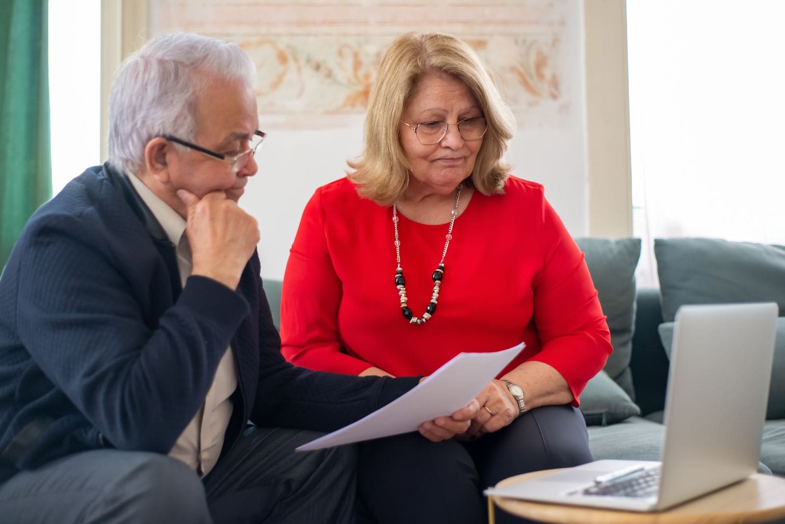 Older couple on a sofa reviewing paperwork beside a laptop